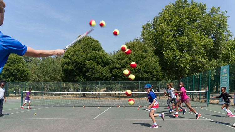 An instructor hitting tennis balls towards a group of children with their rackets