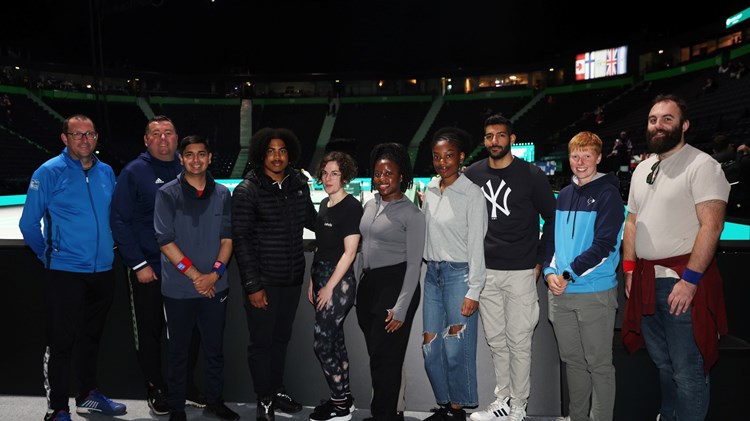 A group of young people stand against a barrier, with a large area of arena seating visible behind them