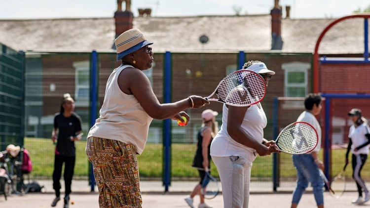 An image of a group of ladies holding tennis rackets and balls on a concrete tennis court group session