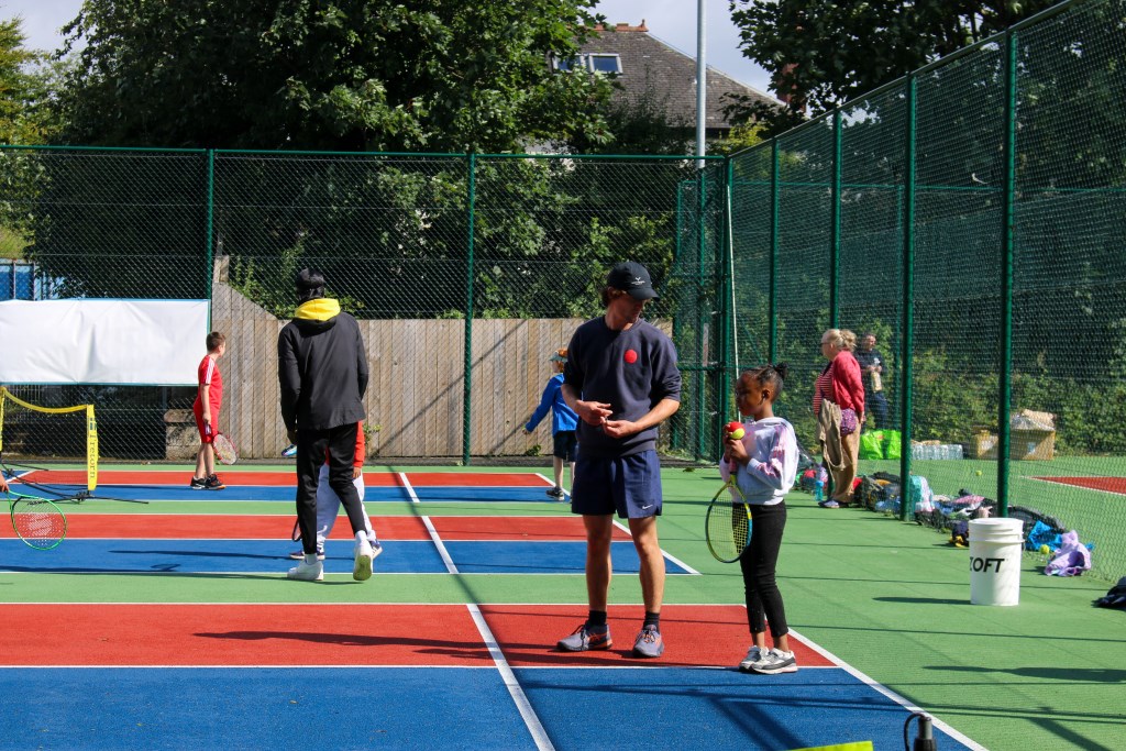 Tennis coach speaking with child on outdoor tennis court. Child is holding racket and ball. 
