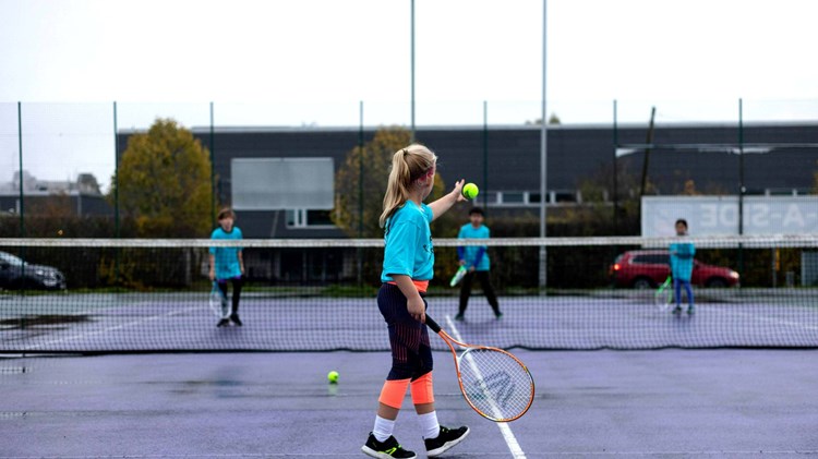 A young girl drops a tennis ball and preapres to hit it over a tennis net in the direction of three other children stood on the other side of the net