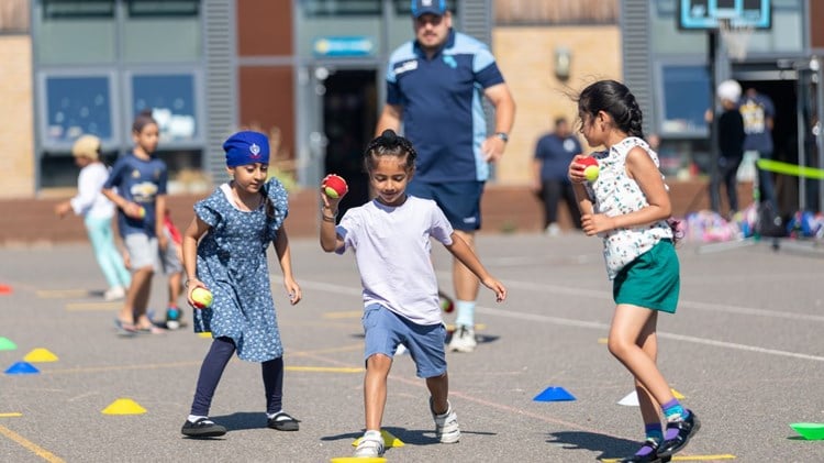 three children playing with tennis balls on a outdoor play area with a coach in the background