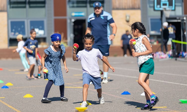 three children playing with tennis balls on a outdoor play area with a coach in the background