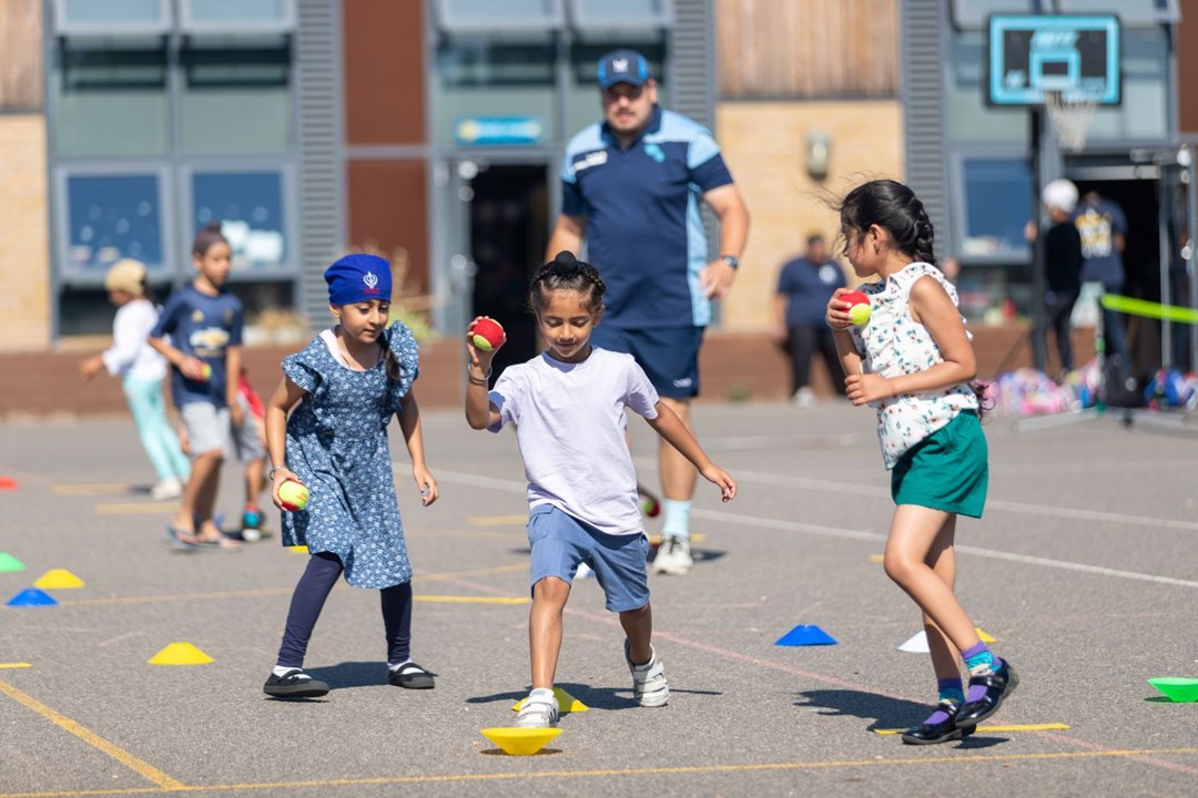 three children playing with tennis balls on a outdoor play area with a coach in the background