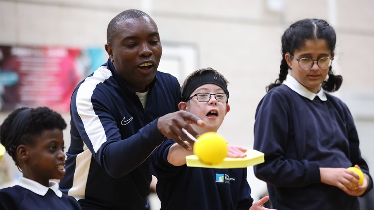 A coach standing behind a young boy, helping him hit a soft ball with a padel