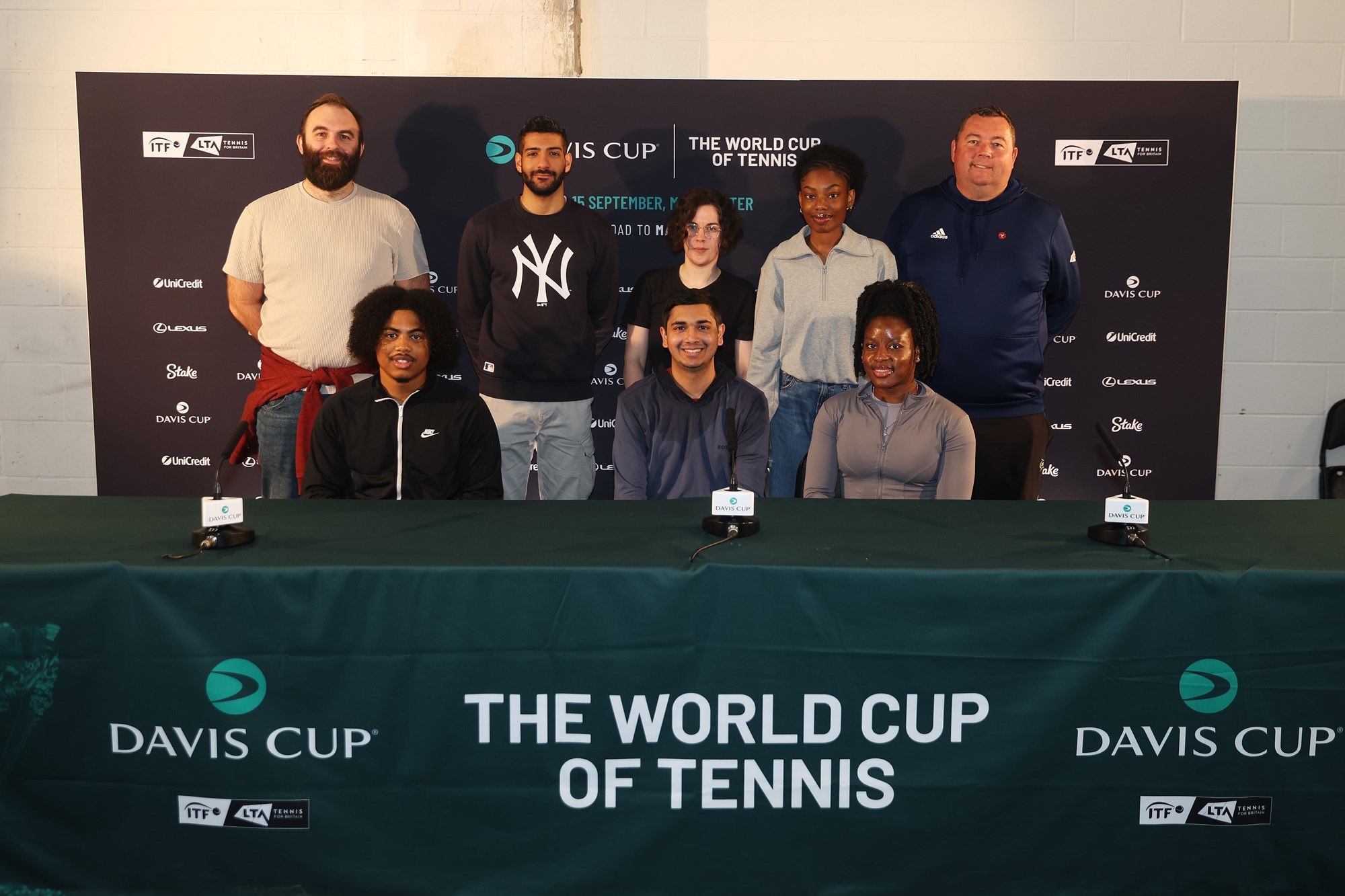 5 people standing and 3 people sitty at the Davis Cup press conference table with the sponsor board behind