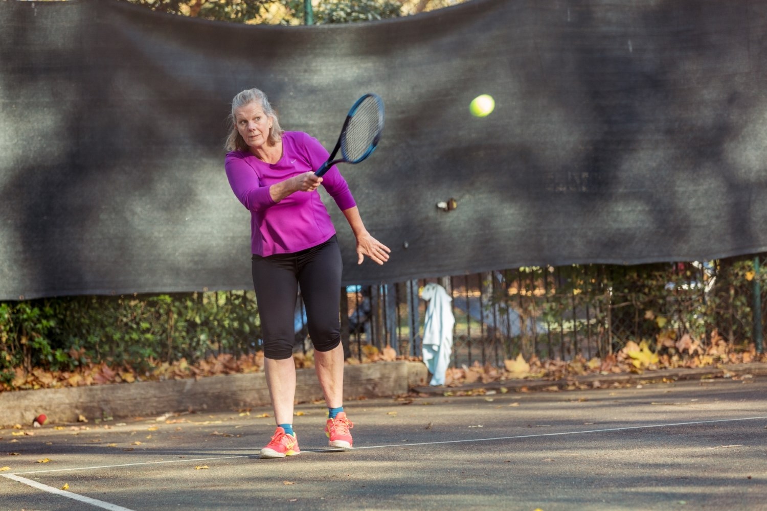 A woman is stood on a concrete tennis court, wearing a purple long-sleeved top and black knee-length bottoms hold a blue tennis racket, with a tennis ball mid-air after being hit.