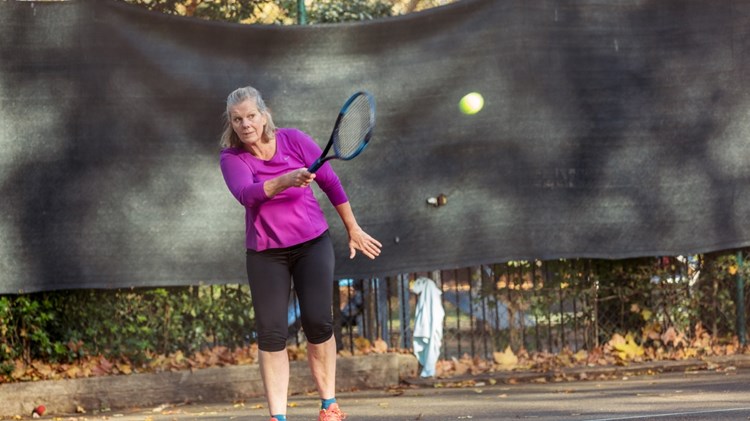 A woman is stood on a concrete tennis court, wearing a purple long-sleeved top and black knee-length bottoms hold a blue tennis racket, with a tennis ball mid-air after being hit.