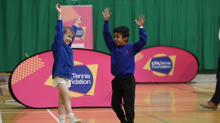 Two children smile with their arms in the air in a school sports hall. On the ground behind them is a pink banner wuth the words, 'Tennis Foundation' visible