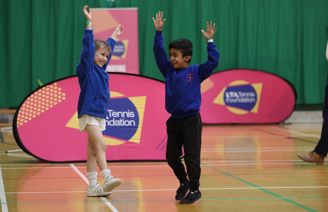 Two children smile with their arms in the air in a school sports hall. On the ground behind them is a pink banner wuth the words, 'Tennis Foundation' visible