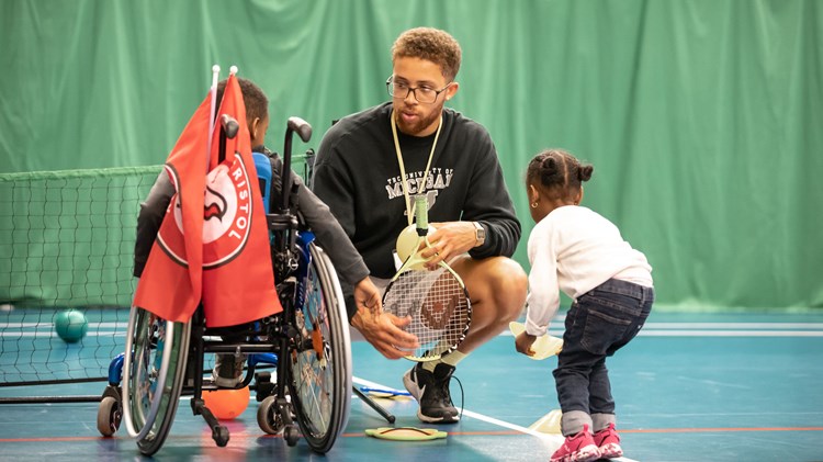 A man crouches to talk to two young children. He is holding a tennis racket, while the child on his right facing him is in a wheelchair, and the child on his left picks up a cone
