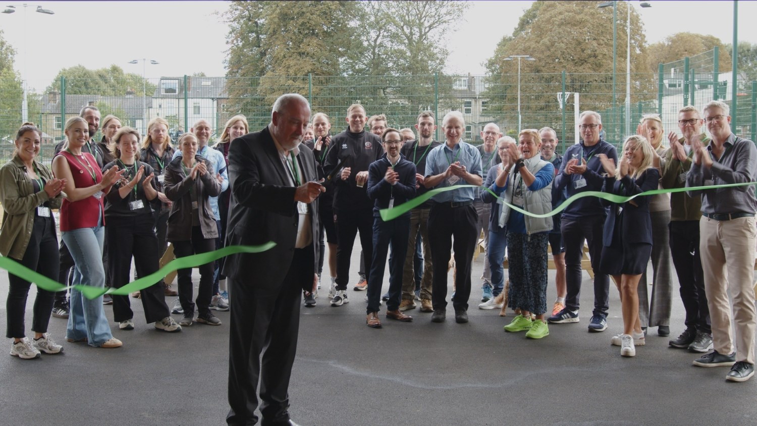 A man holds a pair of scissors having just cut a green ribbon, which is now in two pieces still waving in the air. Behind the man, a large group of people smile and clap.