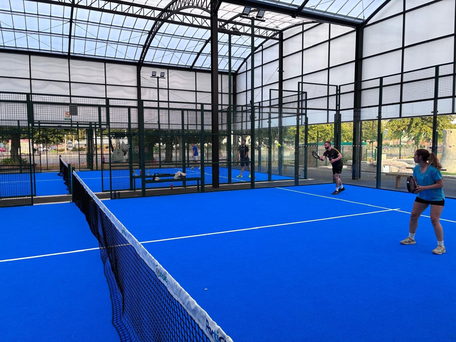 Two people stand at the back of an enclosed padel court, the court surface of which is blue in colour. There is another court in the background, and both courts are covered by a large white canopy supported by black supports.