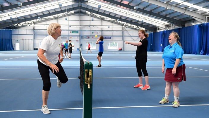 Two young players on one side of the net, laughing as their coach displays a move at the other side