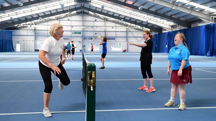 Two young players on one side of the net, laughing as their coach displays a move at the other side
