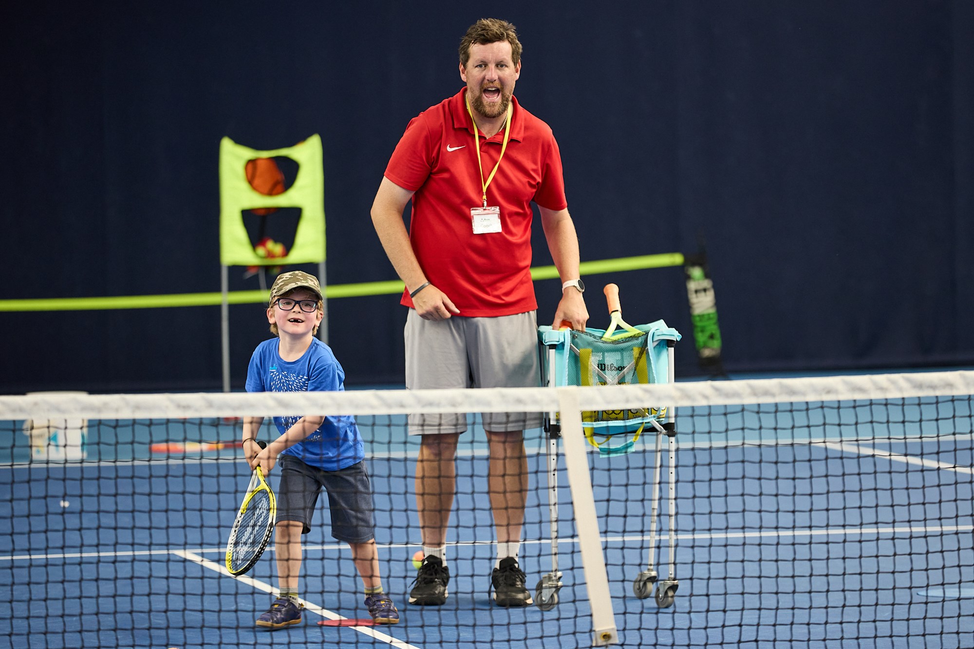 Tennis coach and child standing at tennis net smiling. Child is holding a racket