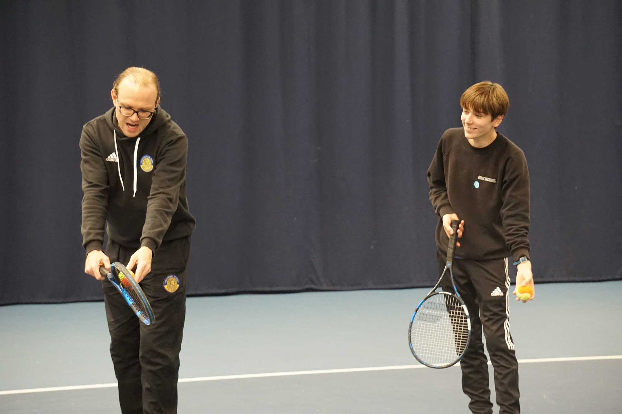 An older boy and a man both stand with tennis rackets and a ball each. The boy watches as the man demonstrates how to serve.