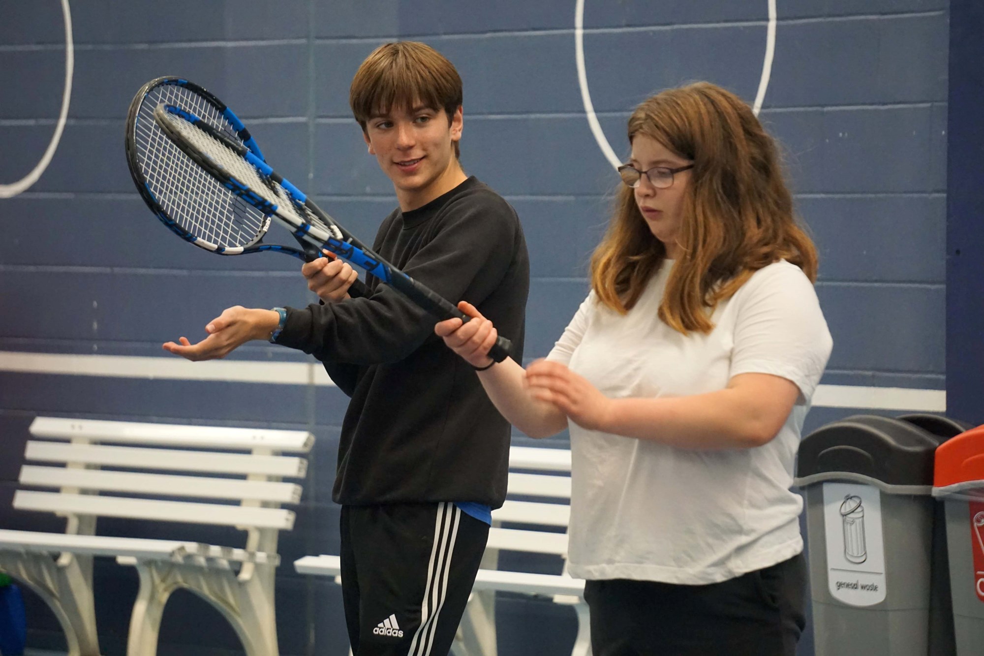 Two young people, one boy and one girl, both stand holding tennis rackets.