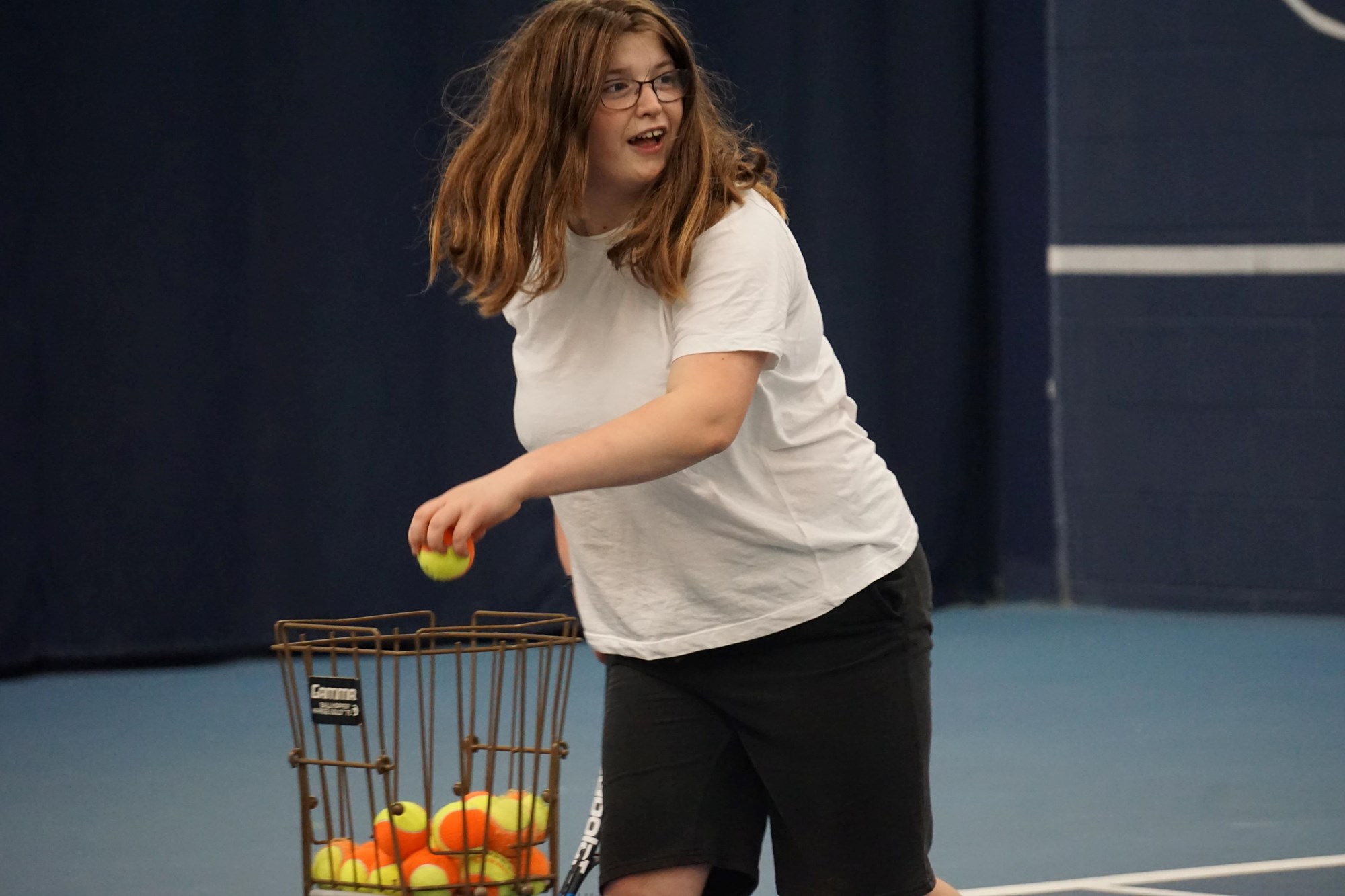 A young woman picks a tennis ball out of a wire crate that holds more tennis balls