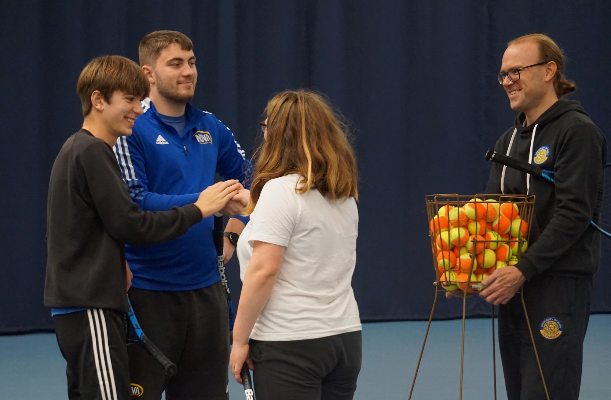Four people stand together. One man holds a crate of tennis balls, while the other three stand in a group with their hands in the middle