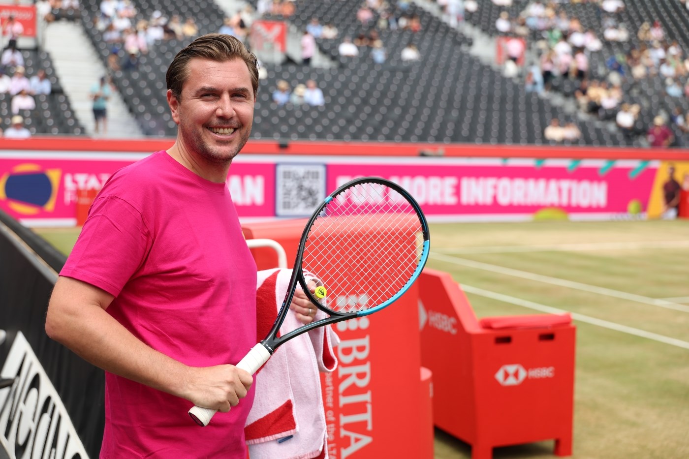 A man in a pink LTA Tennis Foundation t-shirt smiling and holding a racket about to head on court at The Queen's Club