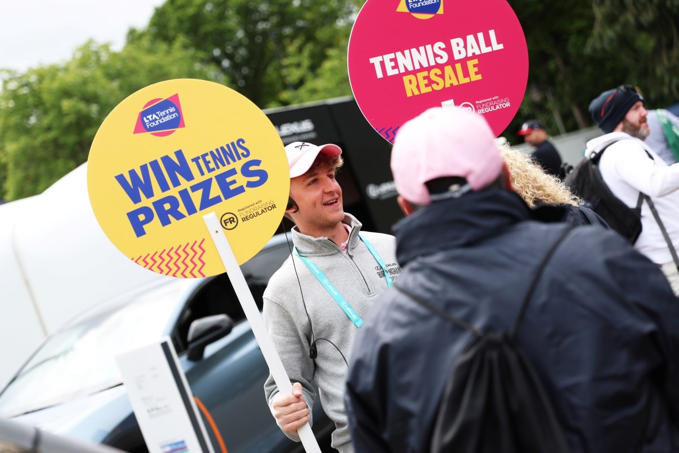 A male staff member holding up two placards advertising the LTA Tennis Foundation prize draw and tennis ball resale