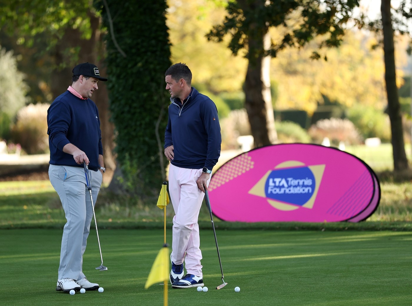Two men, both wearing dark blue shirts and grey trousers, stand with golf balls at their feet and each holding a putter in their hand. They stand on a large area of grass, and behind them , a large banner with the words, 'LTA tennis Foundation' is visible.