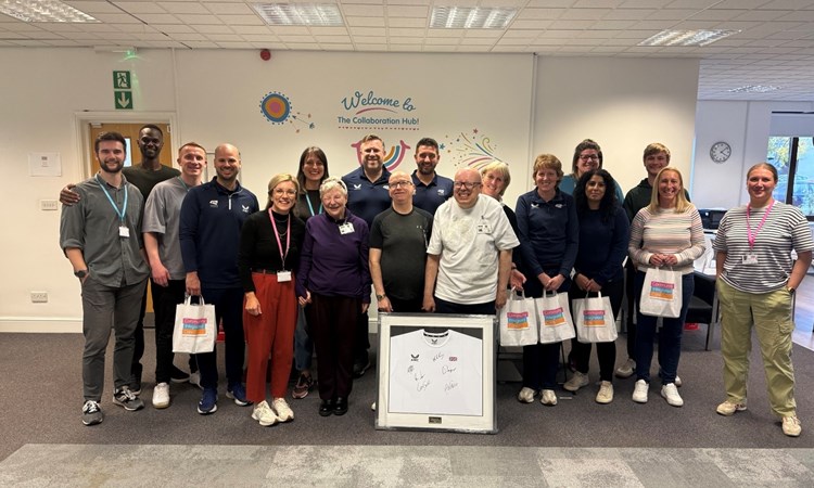 A group of people standing together looking at the camera for a group photo. In the middle is a framed tennis shirt signed by British players 