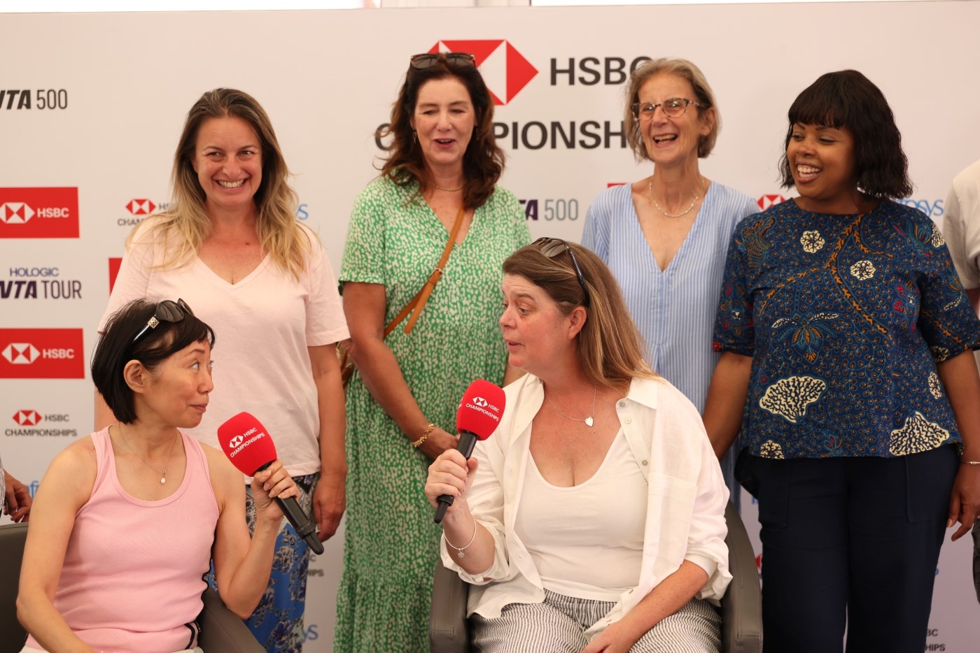 Two women sitting down speaking intomicrophones with four women standing behind, looking on and smiling