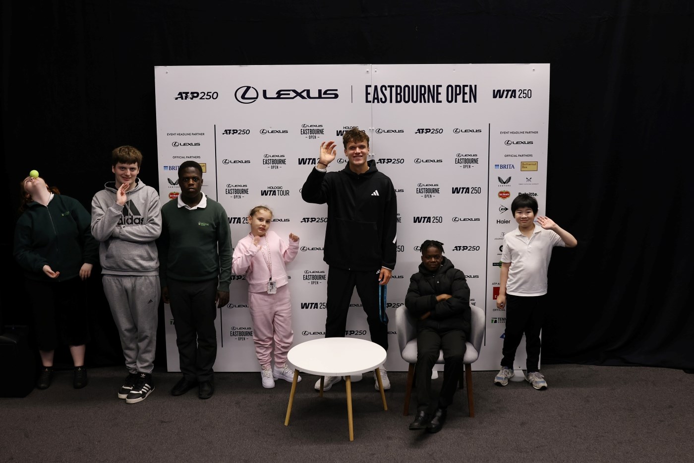 A line up of six children standing and one sitting in front of a Lexus Eastbourne Open press board smiling and waving