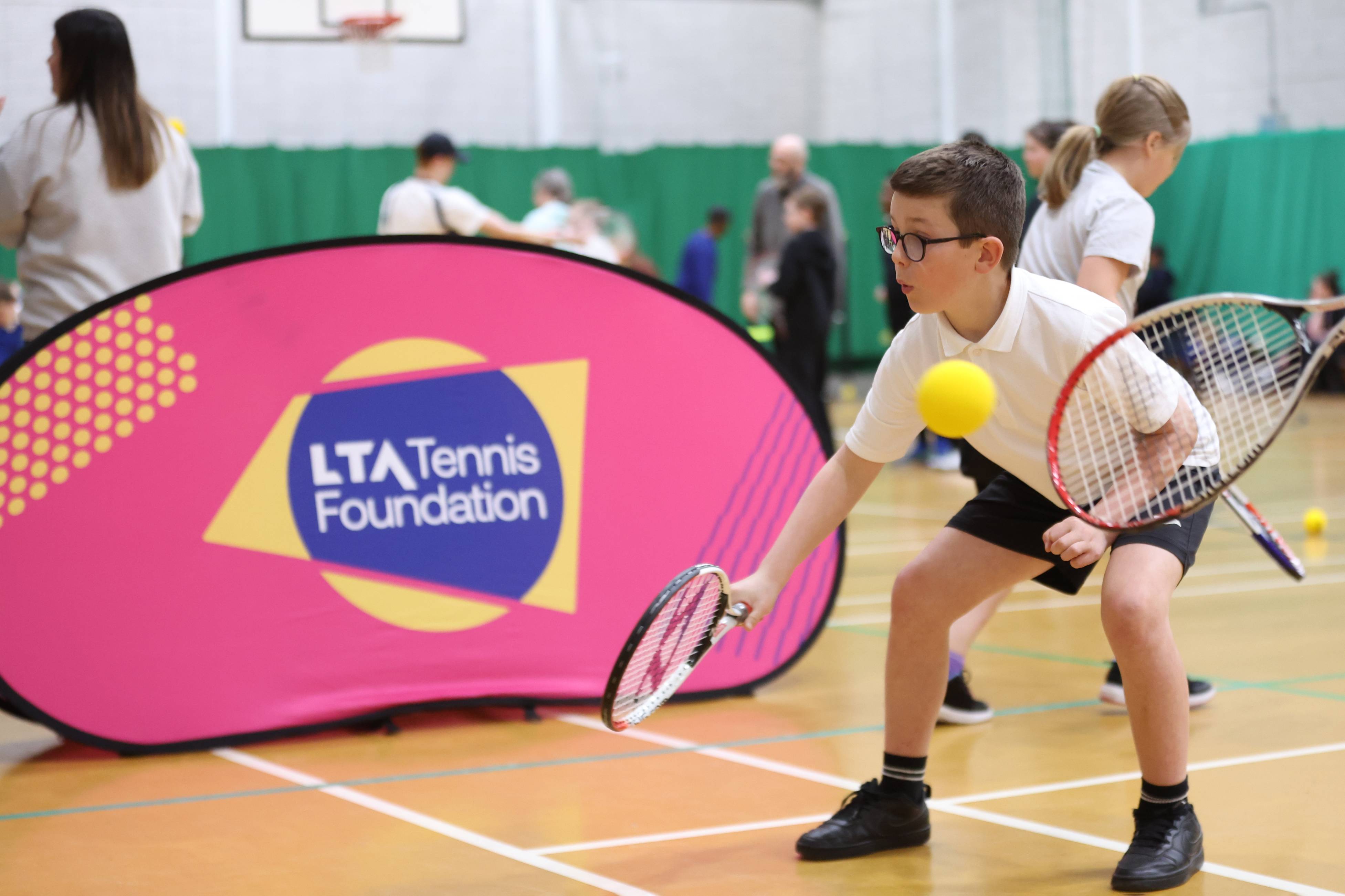 A boy in a white school uniform crouches low whilst holding a tennis racket. Behind him is a large pink banner with the words 'LTA tennis Foundation' on it, and in the foreground, a yellow sponge ball and tennis racket held by another person are visible.