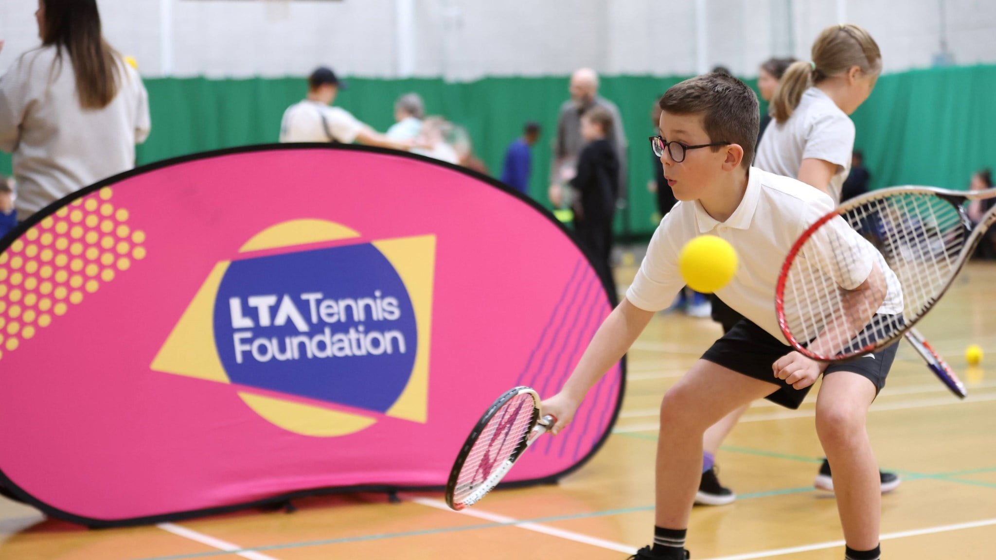 A boy in a white school uniform crouches low whilst holding a tennis racket. Behind him is a large pink banner with the words 'LTA tennis Foundation' on it, and in the foreground, a yellow sponge ball and tennis racket held by another person are visible.