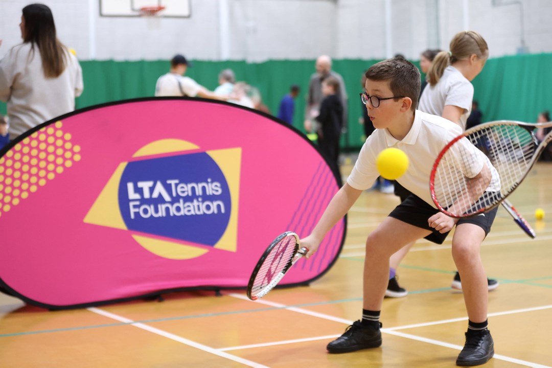 A boy in a white school uniform crouches low whilst holding a tennis racket. Behind him is a large pink banner with the words 'LTA tennis Foundation' on it, and in the foreground, a yellow sponge ball and tennis racket held by another person are visible.