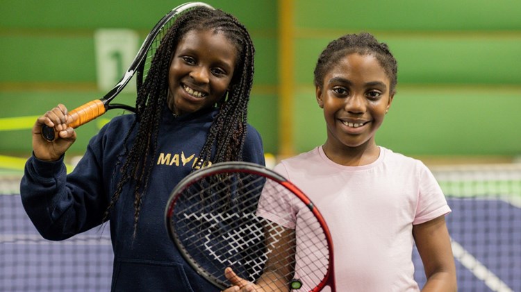 Two girls stand side-by-side smiling and holding tennis rackets, in front of a tennis net