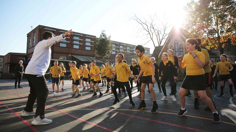 A large group of schoolchildren stand outside on a playground with their knees slightly bent as they watch a man stood in front of them giving instructions