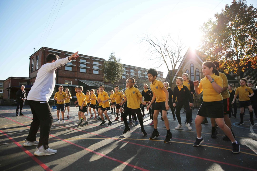 A large group of schoolchildren stand outside on a playground with their knees slightly bent as they watch a man stood in front of them giving instructions