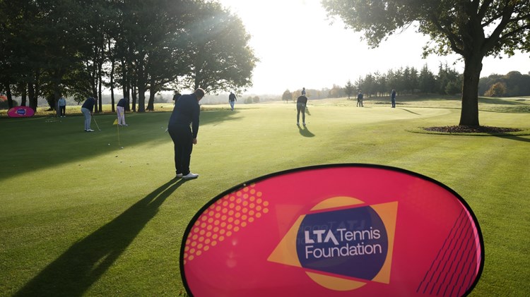 A golf green with an LTA Tennis Foundation banner in the foreground, and people in the background