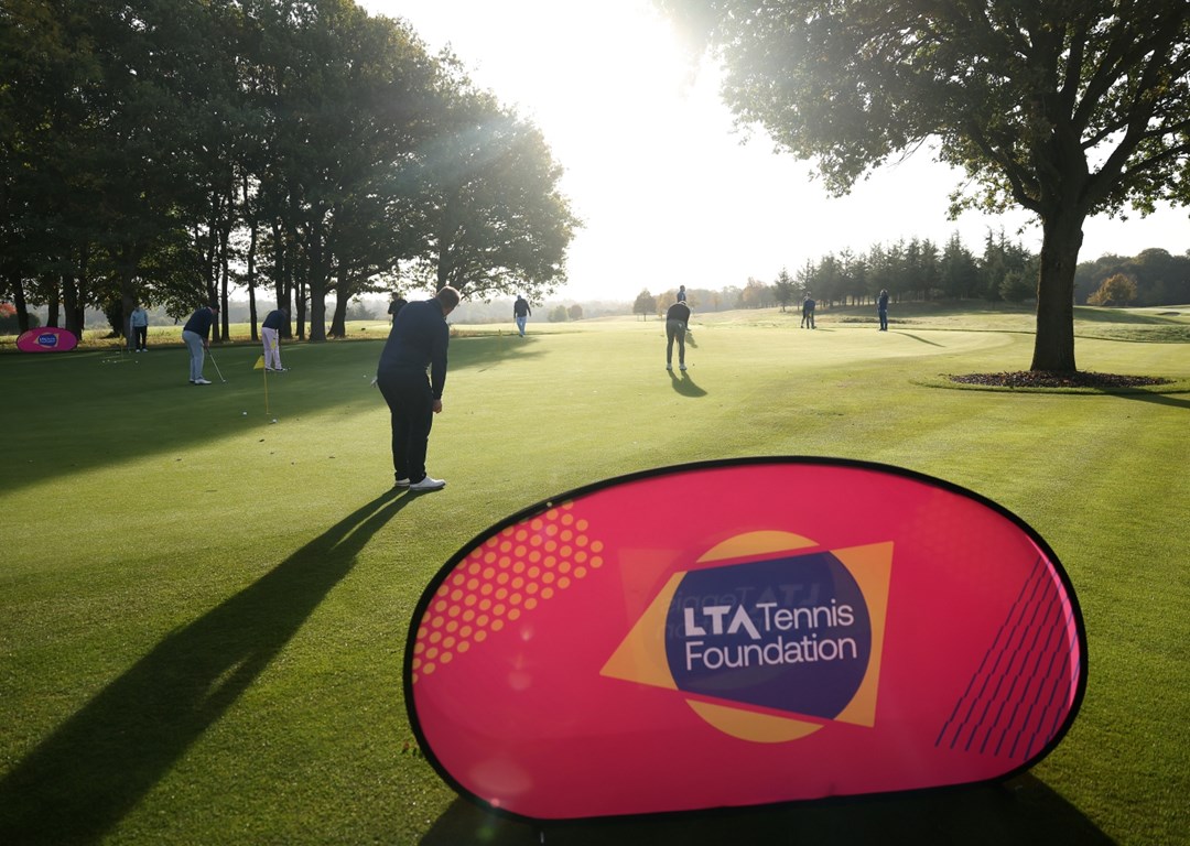 A golf green with an LTA Tennis Foundation banner in the foreground, and people in the background