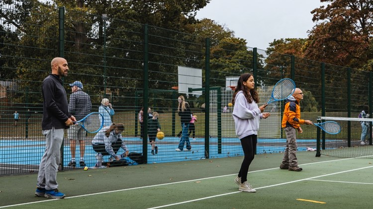 Three peopl two adult men and a young girl, each hold blue tennis racets in theire hands, while standing on a green tennis court. There is fencing behind them, spearting the tennis court from an area where adults and children are bouncing balls, and basketball hoops can be seen
