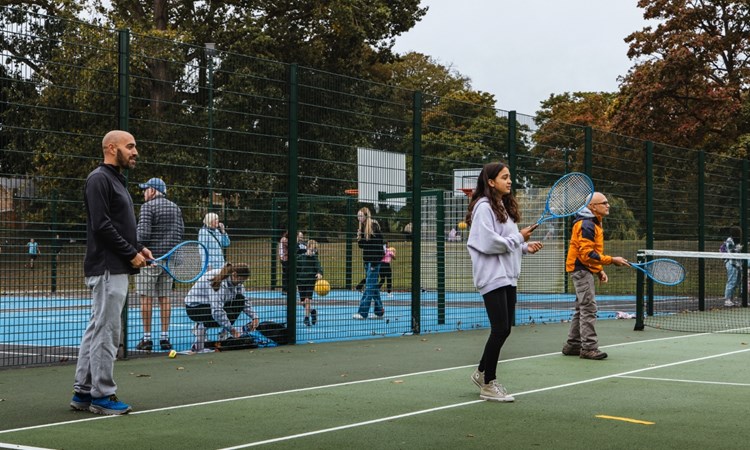Three peopl two adult men and a young girl, each hold blue tennis racets in theire hands, while standing on a green tennis court. There is fencing behind them, spearting the tennis court from an area where adults and children are bouncing balls, and basketball hoops can be seen