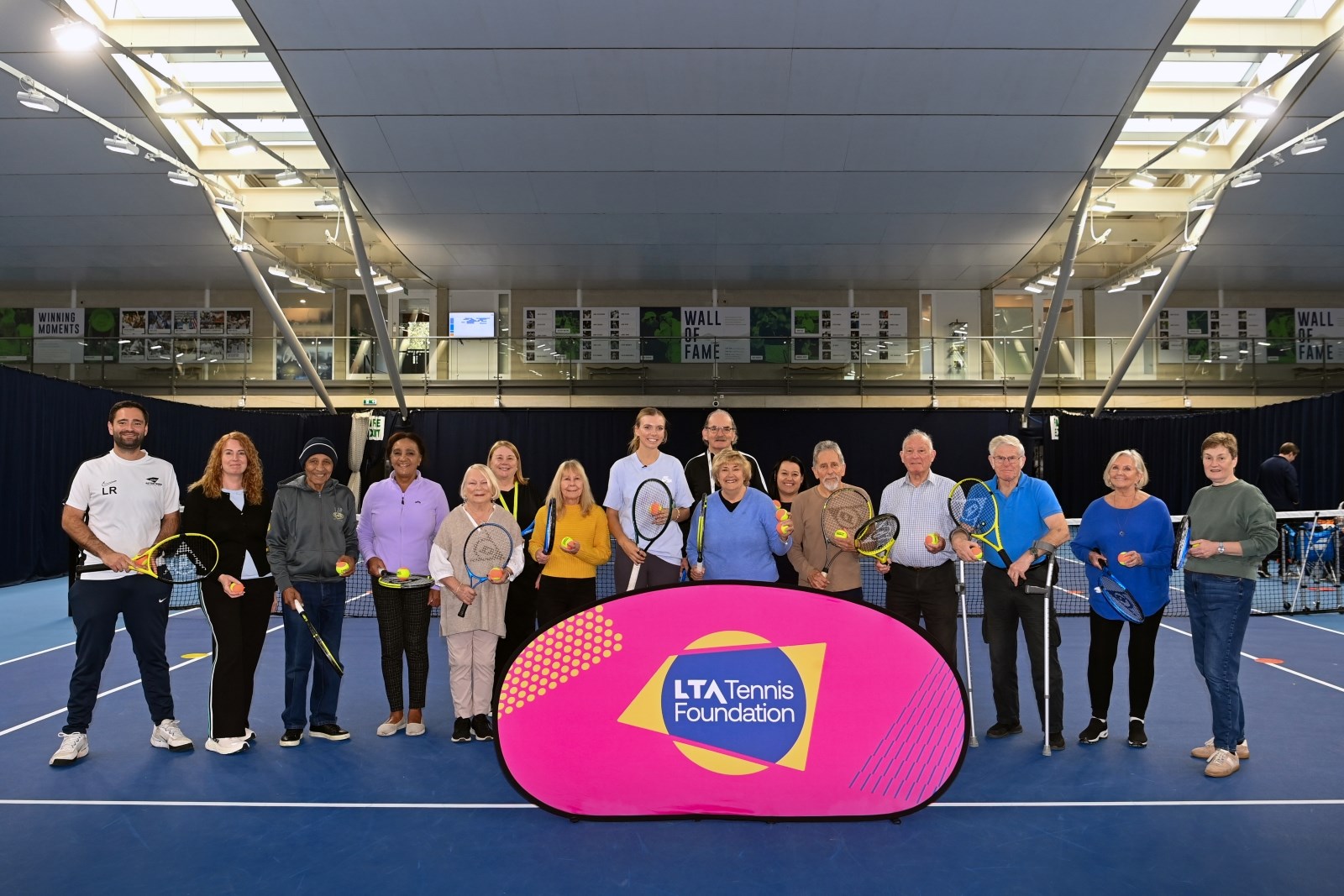 A group of elderley people, with a young woman in the middle of the grop and a younger man at the far left of the group, stand on a tennis court holding tennis rackets, with a pink sign in front of them with the words, 'LTA Tennis Foundation' on it.