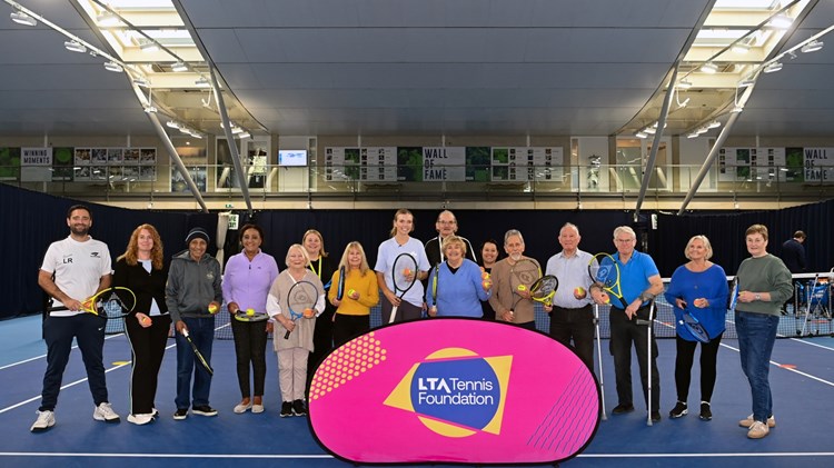 A group of elderley people, with a young woman in the middle of the grop and a younger man at the far left of the group, stand on a tennis court holding tennis rackets, with a pink sign in front of them with the words, 'LTA Tennis Foundation' on it.