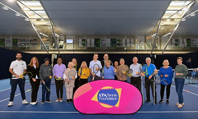 A group of elderley people, with a young woman in the middle of the grop and a younger man at the far left of the group, stand on a tennis court holding tennis rackets, with a pink sign in front of them with the words, 'LTA Tennis Foundation' on it.