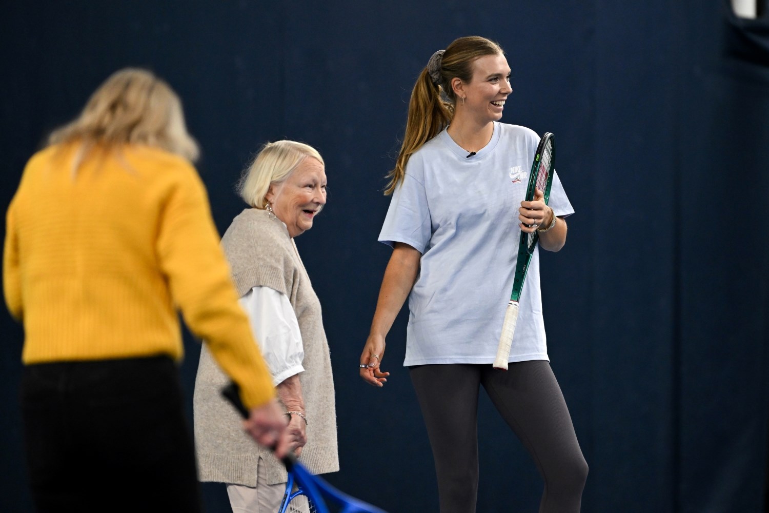 Two women, one elderly and one younger, stand next to each other, smiling and holding tennis racquets. At the edge of the image in the foreground, a woman wearing a yellow jumper who also holds a tennis racquet has her back turned.