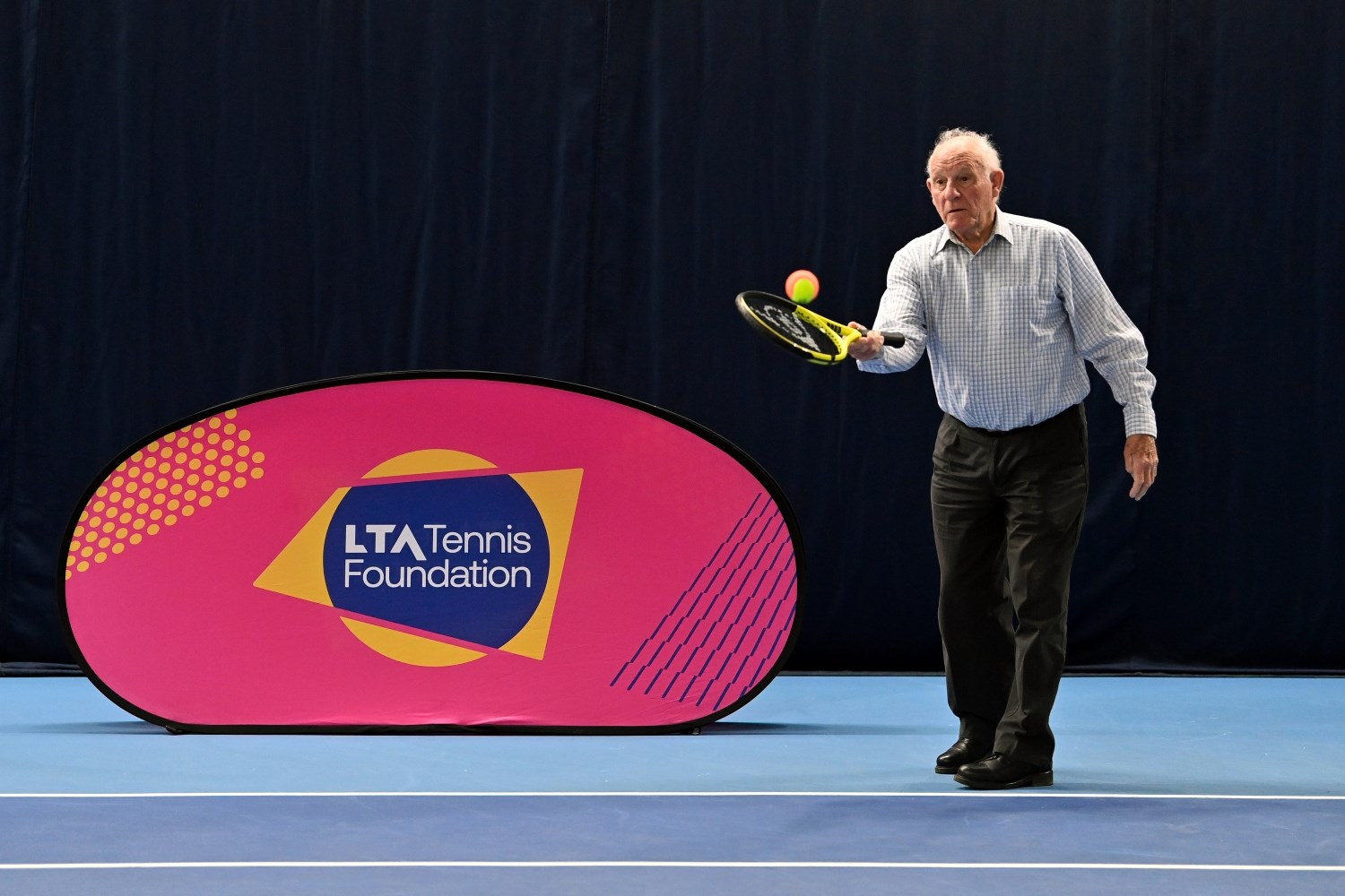 An old man hits a red and yellow tennis ball with a tennis racket. Next to him is a pink sign with the words, 'LTA Tennis Foundation' written on it