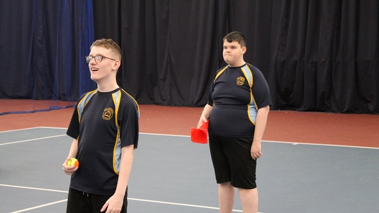 Two students on a tennis court at an indoor centre. One is holding a ball and one is holding a cone ready to play a tennis skills game