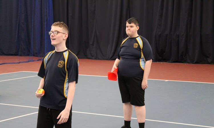 Two students on a tennis court at an indoor centre. One is holding a ball and one is holding a cone ready to play a tennis skills game