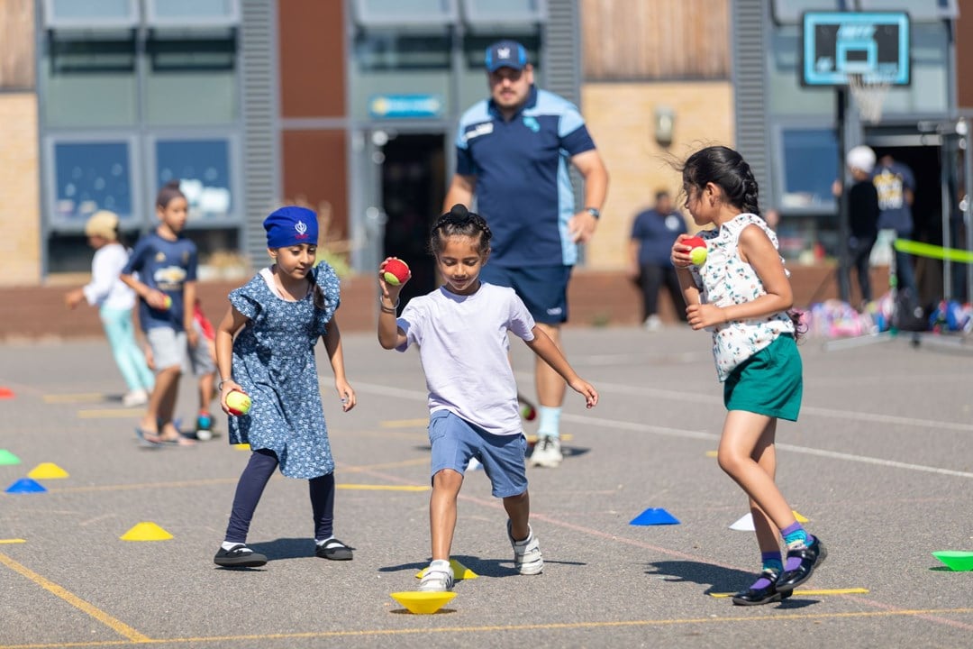 Three young kids playing with tennis balls and cones on the ground while their coach watches on