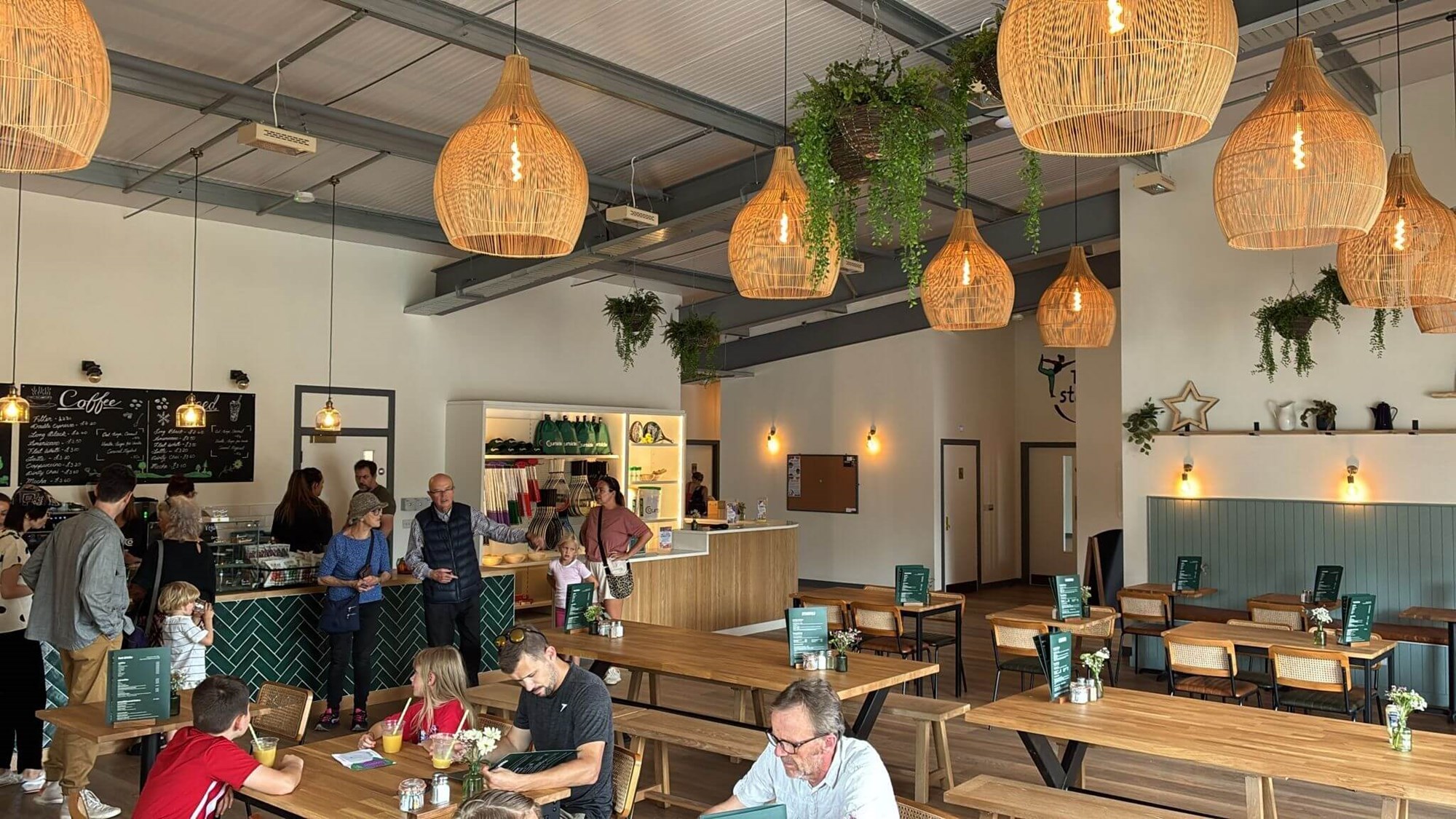 A large open plan room filled with wooden tables and benches. Hangin from the ceiling are lights with large shades on them. At the back of the room, people queue at a counter stacked with food offerings, and on the back wall a blackboard with 'Coffee' written in large letters displays menu options