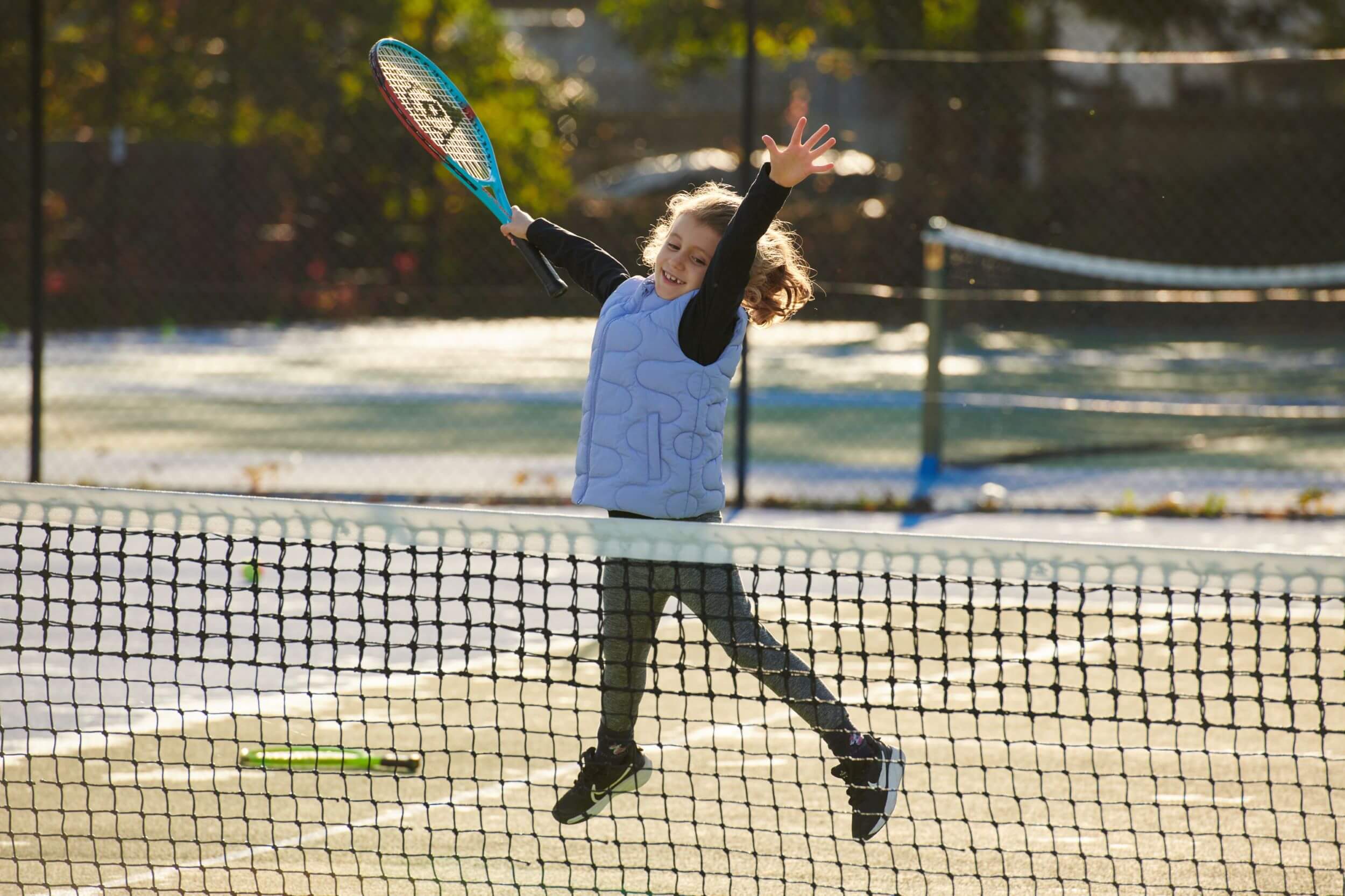 A young girl does a star jump while smiling behind a tennis net with a racket in hand